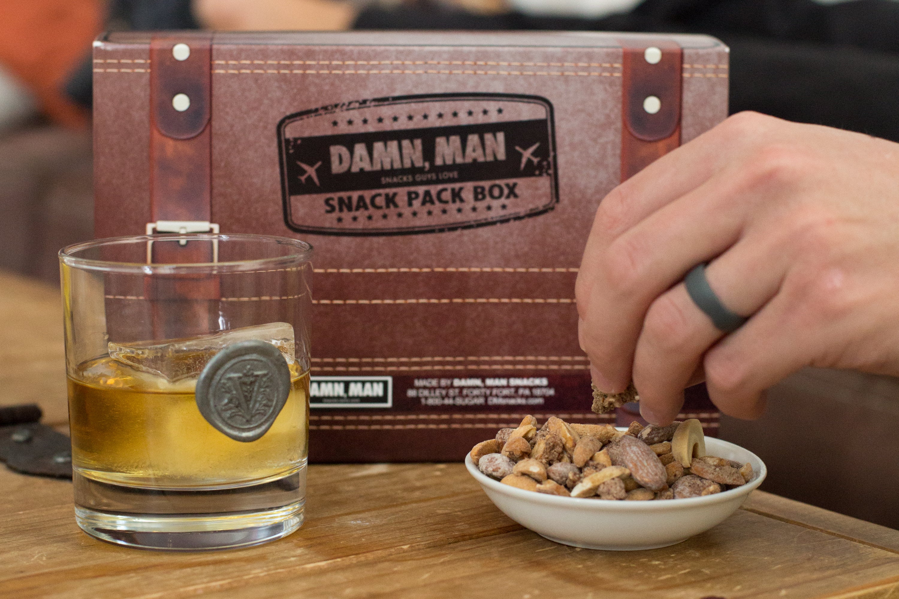 Man grabbing mixed nuts from bowl beside a glass of whiskey and Damn, Man Snack Pack Box in background. Healthy high-protein snack gift box. Perfect Fathers Day Gift Box
