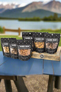Build Your Own 6-Pack Box of Mixed Nuts by Damn, Man Gift Snacks displayed outdoors with scenic mountain background.