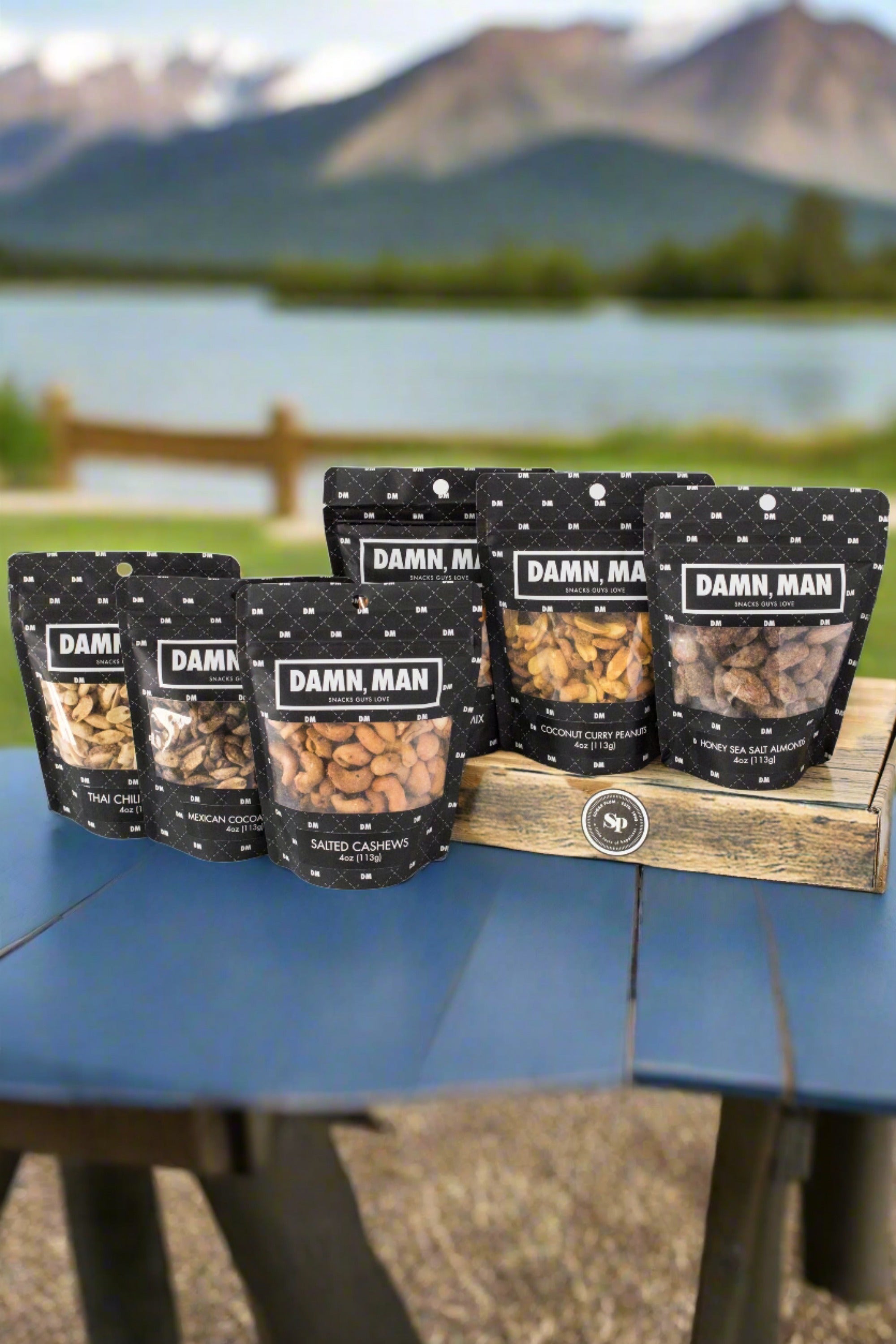 Build Your Own 6-Pack Box of Mixed Nuts by Damn, Man Gift Snacks displayed outdoors with scenic mountain background.