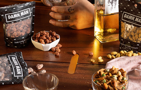 Person holding a glass of whiskey with snacks and 'Damn Man' snack bags on a wooden table.