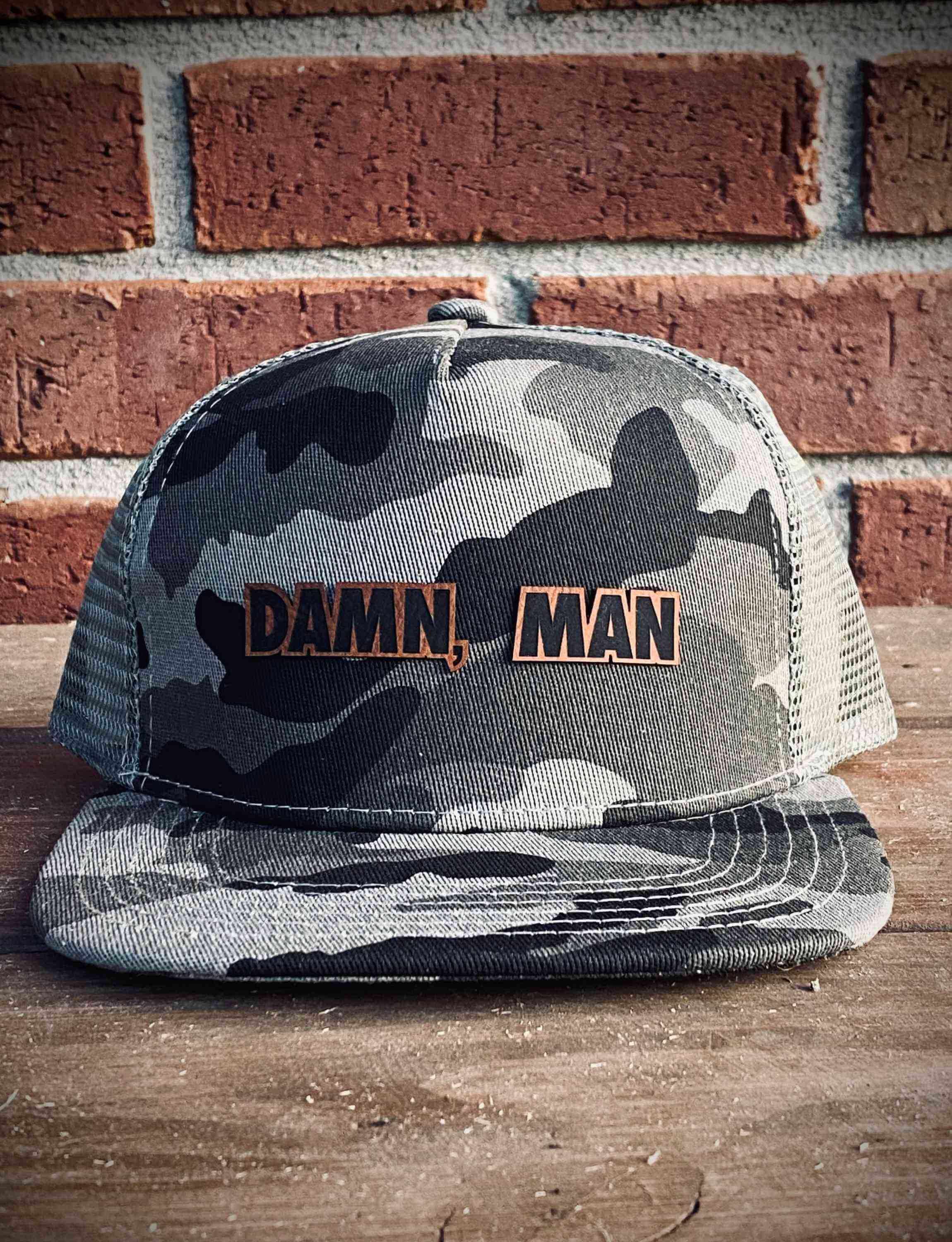 Camo Men's Trucker Hat with Damn, Man text in front of a brick wall background