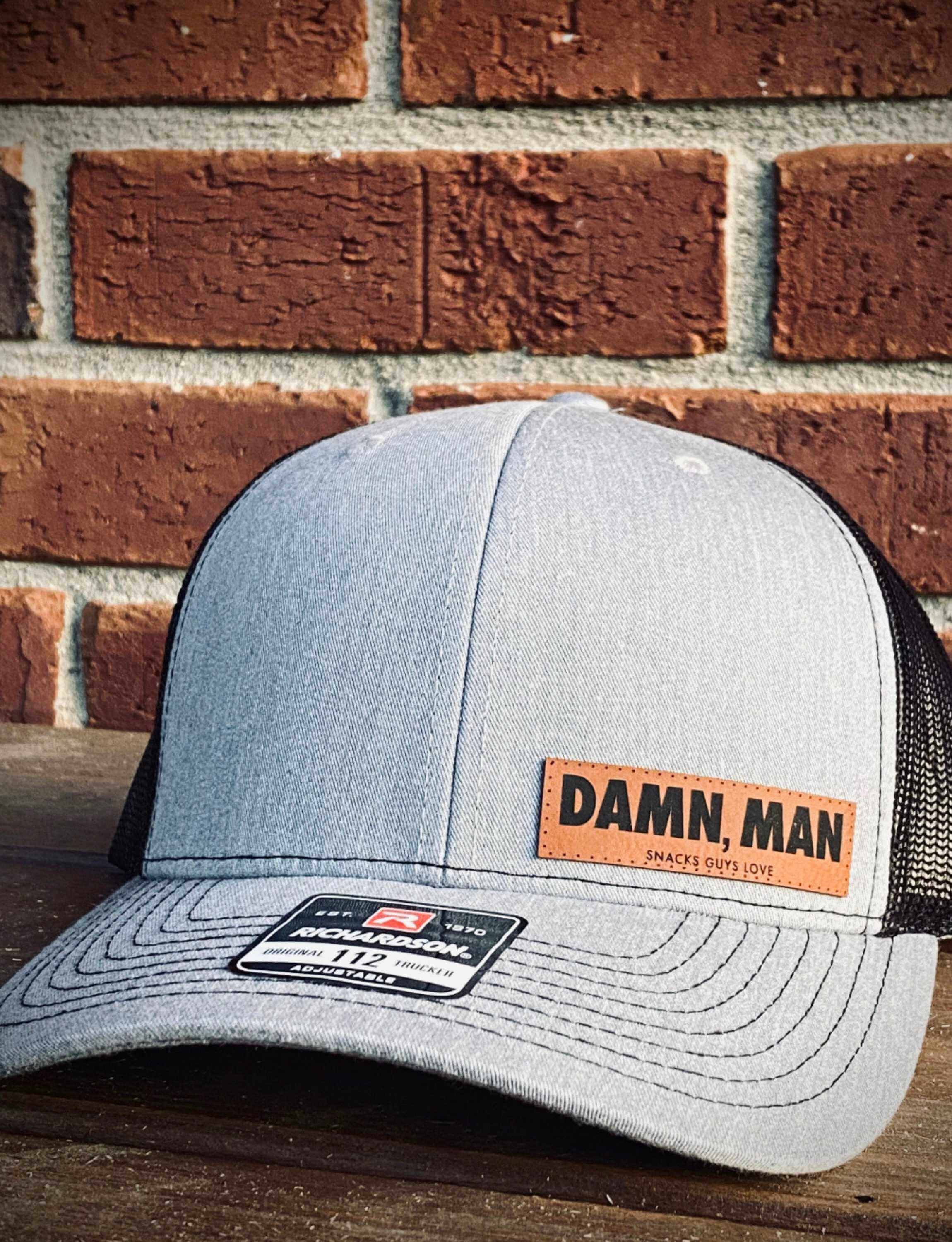 Grey trucker hat with net backing and "Damn, Man" text in front of a brick wall.